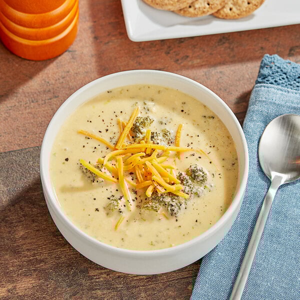 A bowl of Blount Fine Foods broccoli and cheddar cheese soup with cheese and crackers on a white table.
