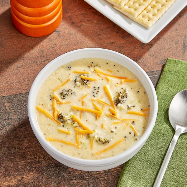 A bowl of Kettle Cuisine Cheddar Broccoli soup with cheese and crackers on a table.