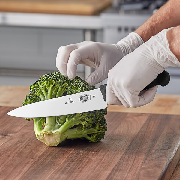 A person using a Victorinox Chef Knife to cut broccoli on a wooden cutting board.