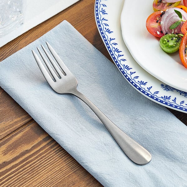 An Acopa stainless steel salad fork on a napkin next to a plate of salad.