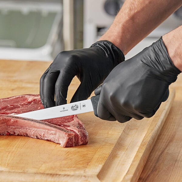 A person wearing black gloves using a Victorinox 6" Wide Stiff Boning Knife to cut meat.