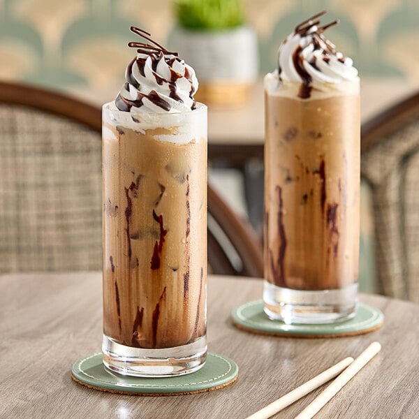 Two glasses of coffee with whipped cream and chocolate drizzle on a table.