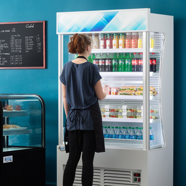 A woman in a hospital cafeteria standing in front of an Avantco white refrigerated air curtain merchandiser with food on display.