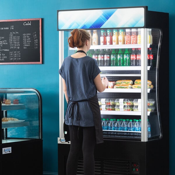 A woman standing in front of a black Avantco refrigerated air curtain merchandiser.