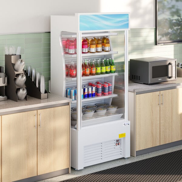 A white refrigerated air curtain merchandiser stocked with drinks and food containers, situated between two countertops in a kitchen or break room setting.