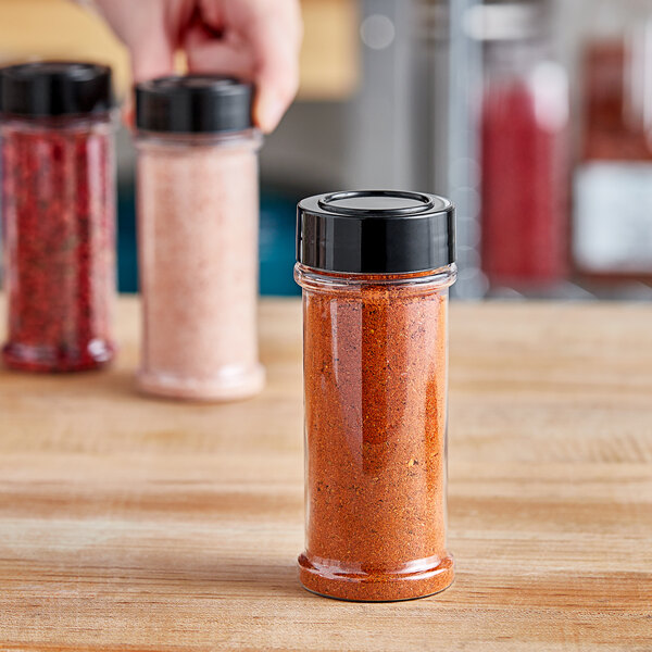 A person holding a round plastic spice container with a black lid filled with red chili pepper.