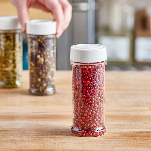 A hand holding a round white plastic spice container filled with red peppercorns.