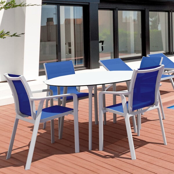 A Siesta Pacific white resin outdoor arm chair with blue sling next to a table on a deck.