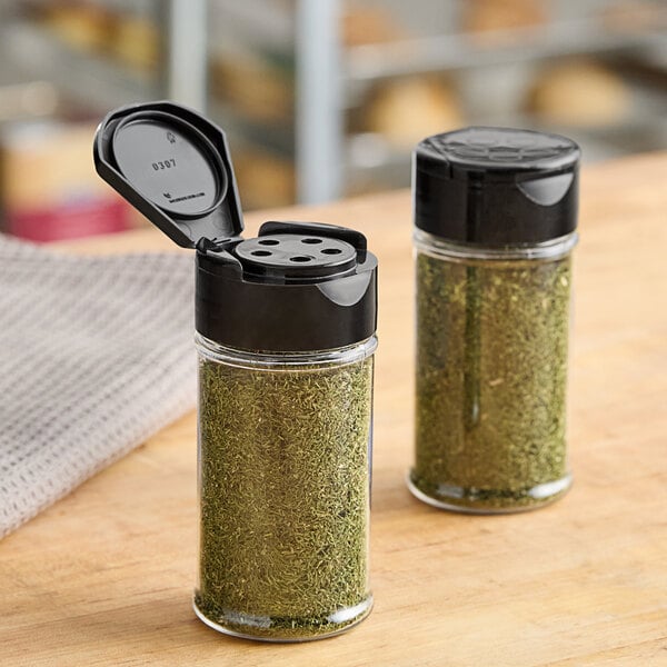 Two round plastic spice containers with black flip and sift lids filled with green herbs.