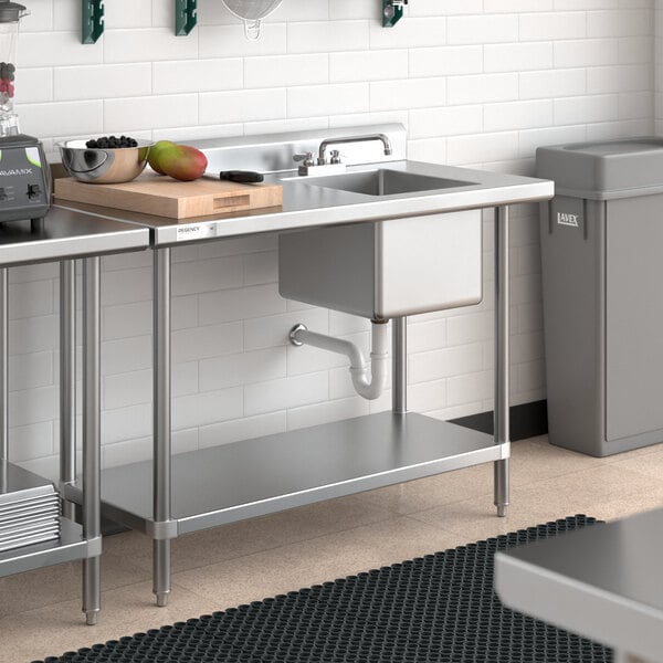 A Regency stainless steel work table with a sink on the right in a kitchen with a sink and a trash can.