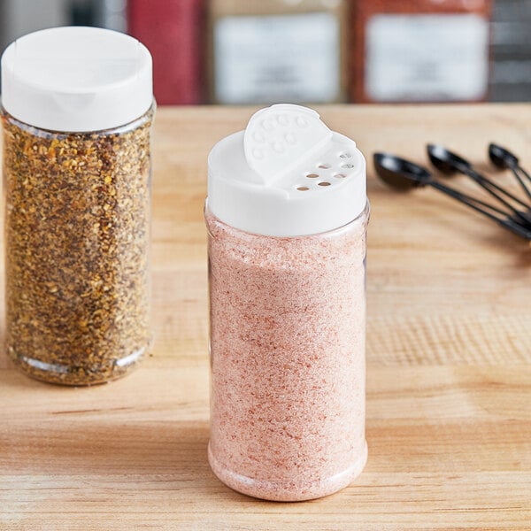 Two 16 oz. round plastic spice containers with white lids filled with seasoning on a wooden table.