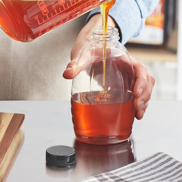 A person pouring honey into a Classic Queenline PET bottle with a black flip top lid on a wood counter.