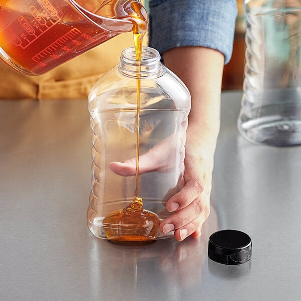 A person pouring honey into a ribbed plastic honey bottle on a kitchen counter.