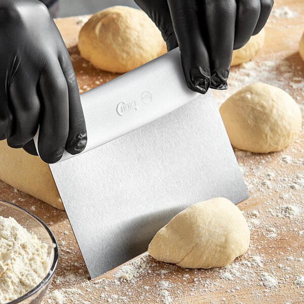 A person using a Choice stainless steel dough cutter with a white handle to cut dough on a table.