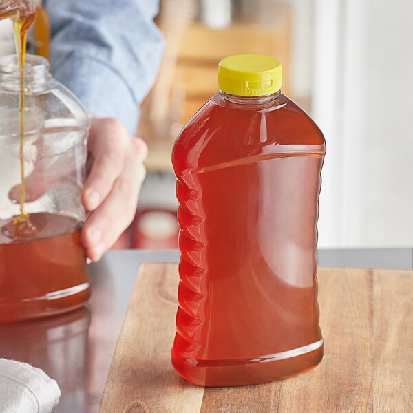 A person pouring honey into a ribbed hourglass PET honey bottle with a yellow plastic flip top lid.