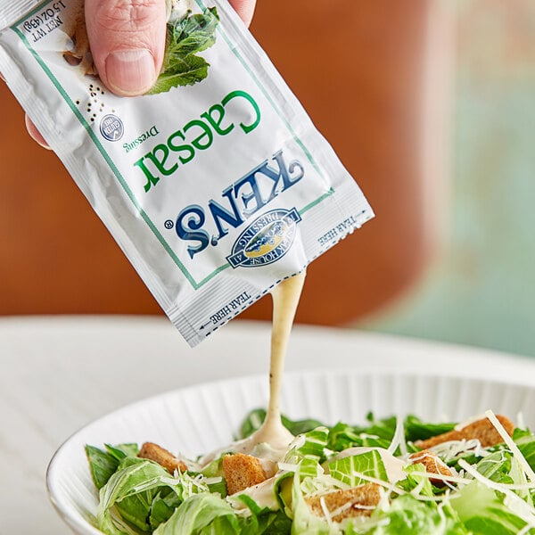 A hand pouring Ken's Caesar dressing onto a bowl of salad.
