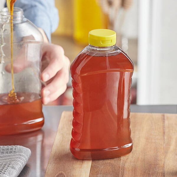 A person pouring honey into a Ribbed Hourglass PET Honey Bottle on a table.