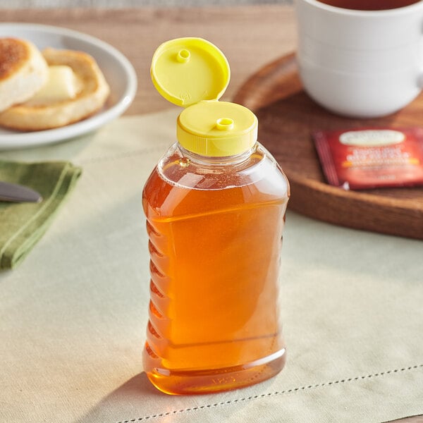 A bottle of honey with a yellow cap on a table next to a cup of tea.