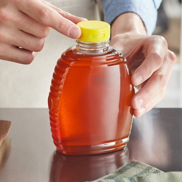 A hand holding a 16 oz. plastic Queenline honey bottle with a yellow flip top lid.