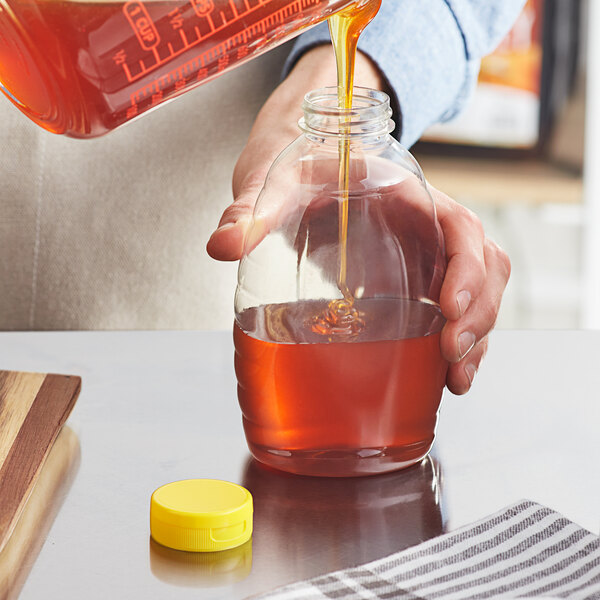 A person pouring honey from a measuring device into a Classic Queenline PET honey bottle with a yellow flip top lid.