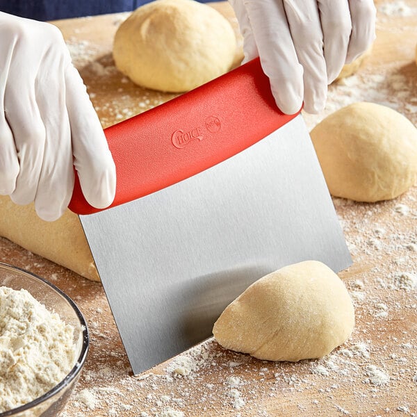 A person using a Choice stainless steel dough cutter with a red handle to cut dough on a surface.