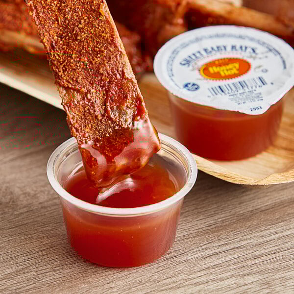A small plastic container of Sweet Baby Ray's Hot Honey Wing Sauce with a piece of meat being dipped into it.