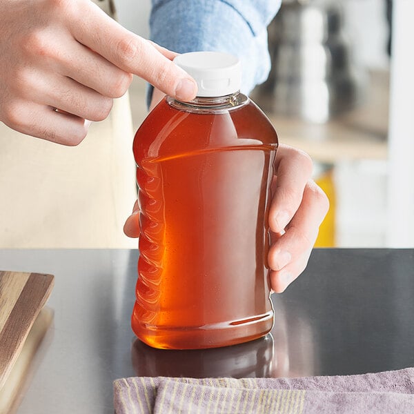 A person pouring honey into a Ribbed Hourglass PET Honey Bottle on a wood surface.