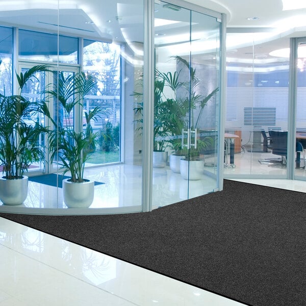 A modern office lobby with large glass walls, several potted plants, and a dark charcoal mat on a shiny tile floor.