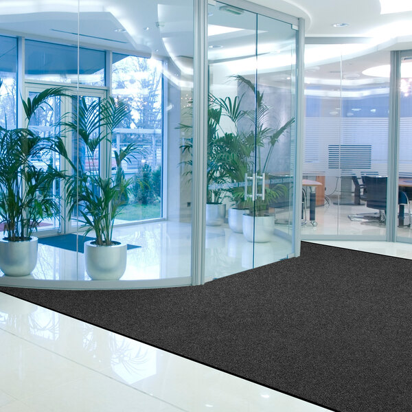 A modern office lobby with large glass walls, several potted plants, and a dark charcoal mat on a shiny tile floor.