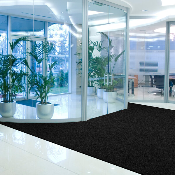 A black rectangular mat with a vinyl border is placed on a shiny white floor in a modern office lobby with glass walls and potted plants.