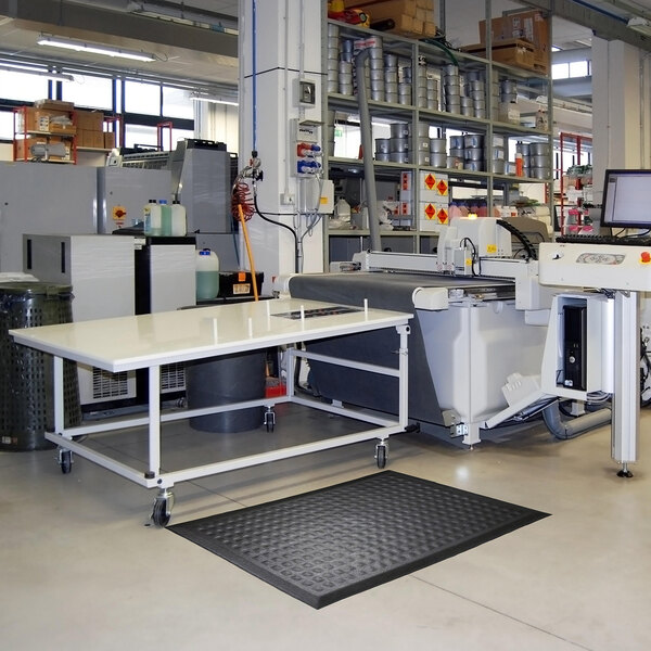 A black anti-fatigue mat with a textured surface and beveled edges is placed on the floor in front of a workstation in an industrial setting.