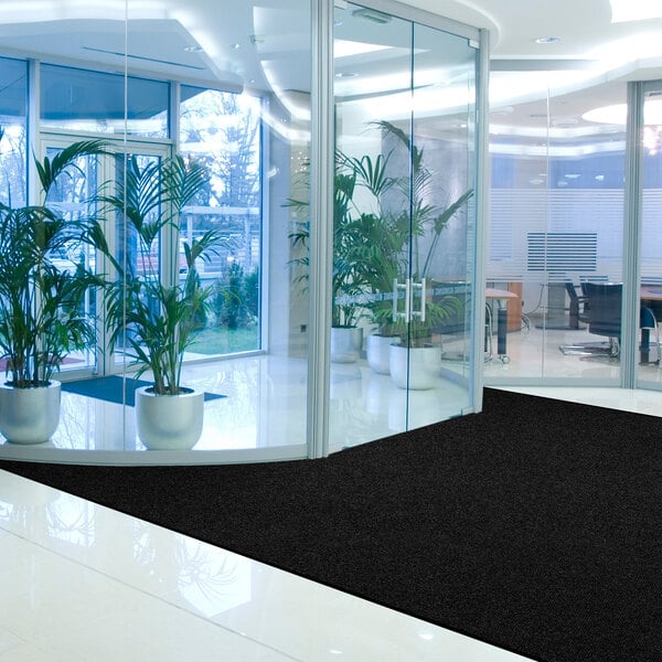 A modern office lobby with large glass walls, several potted plants, and a solid black mat on the shiny floor near the entrance.