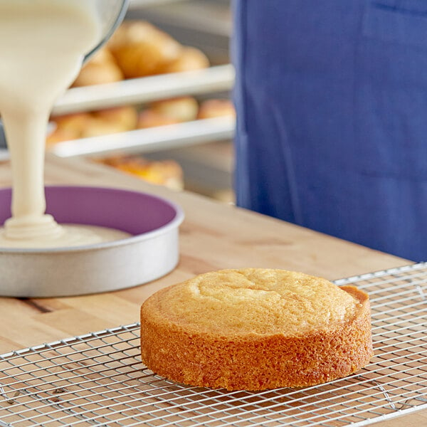 A cake sits on a wire cooling rack. A person is pouring batter into a cake pan in the background.