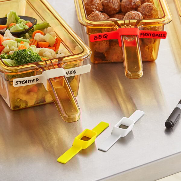 A white rectangular food pan label with a yellow blank label on a table of food in containers.