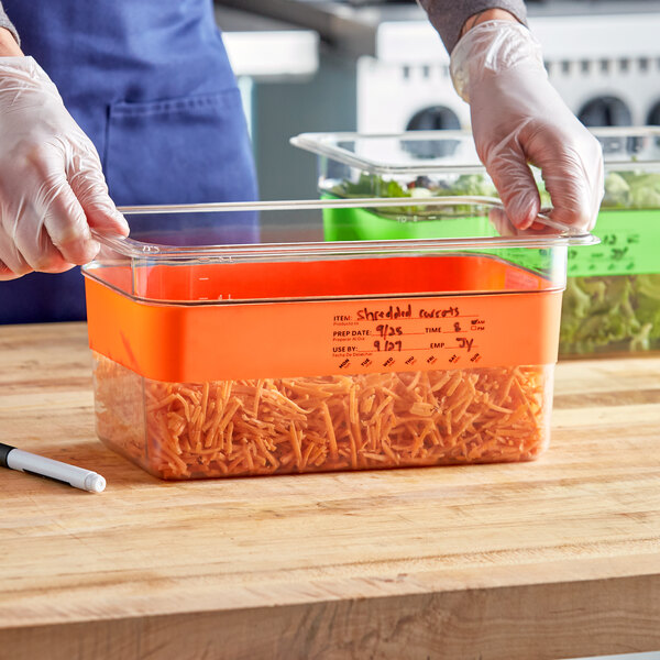 A gloved hand writes on a plastic container of shredded carrots with a Choice Orange Write-On Food Pan Band.