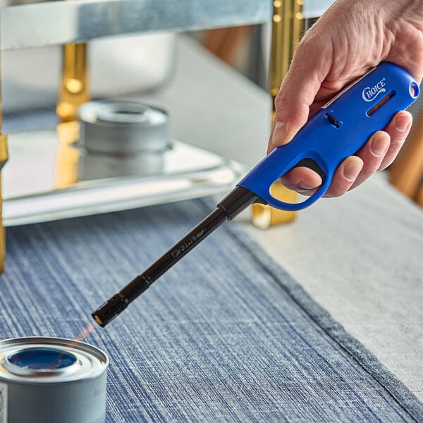 A hand using a blue Choice wind-resistant lighter to light a can of food.