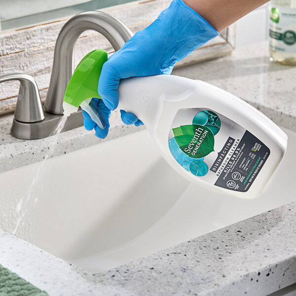 A person wearing blue gloves spraying Seventh Generation Lemongrass Citrus Disinfecting Bathroom Cleaner on a sink.