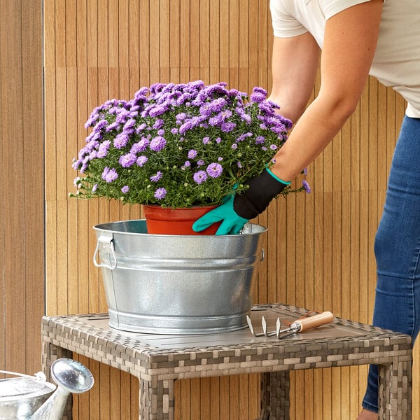 A person wearing gardening gloves holding a potted plant over a Behrens galvanized steel tub.