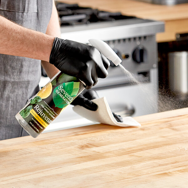 A person wearing black gloves spraying a wood surface with Seventh Generation Yuzu Basil All-Purpose Cleaner.