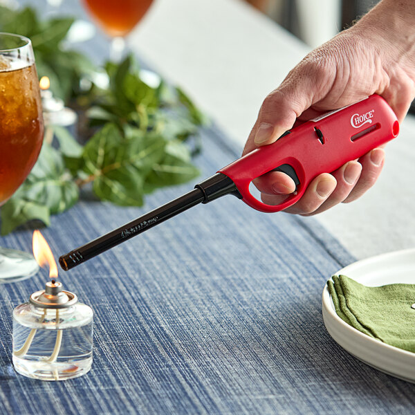 A hand using a red Choice multi-purpose lighter to light a candle on a table with a white bowl and green napkins.