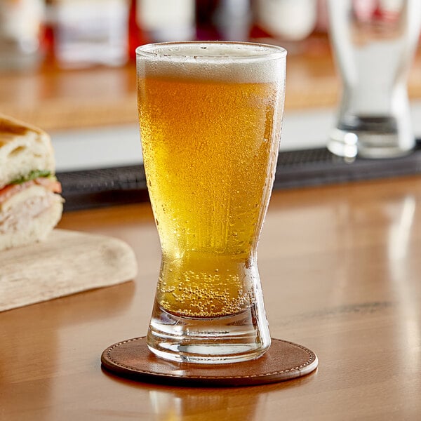 A glass of beer on a coaster on a wooden table next to a sandwich.