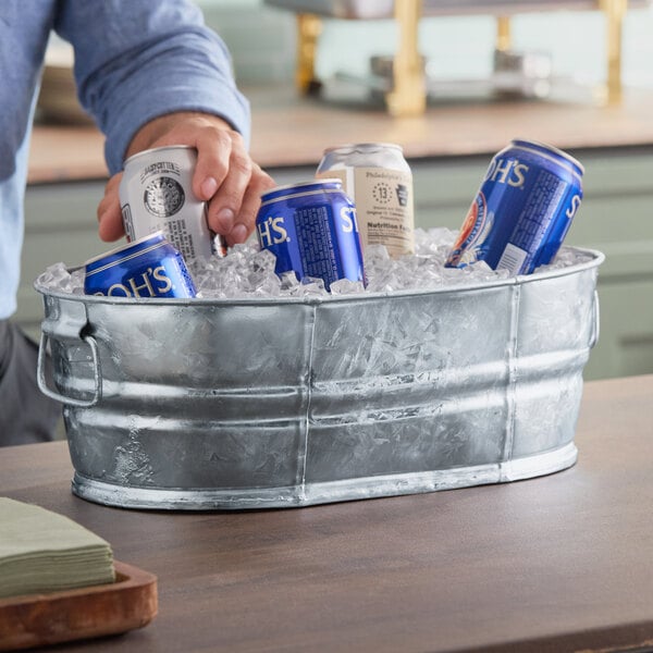 A person holding a blue can of beer in a Behrens steel tub filled with ice.