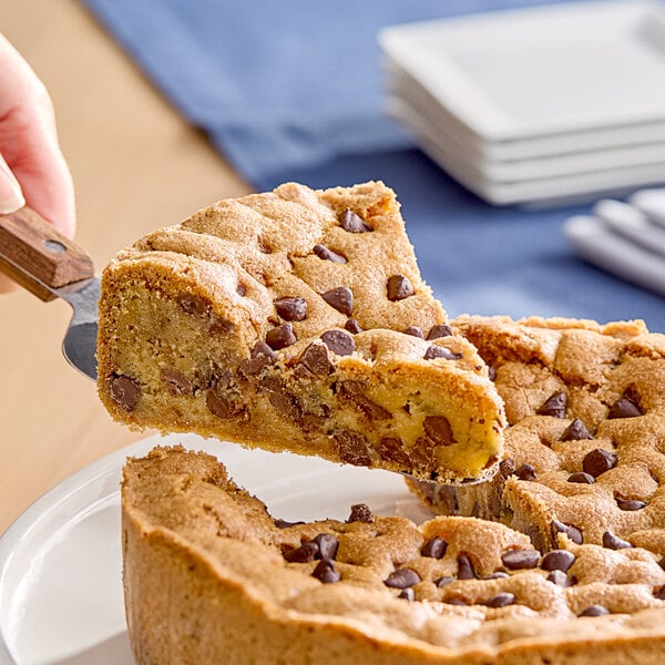 A hand cutting a piece of a chocolate chip cookie cake on a white plate.