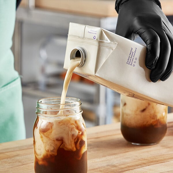 A person in black gloves pouring Minor Figures Barista Organic Oat Milk into a glass jar.