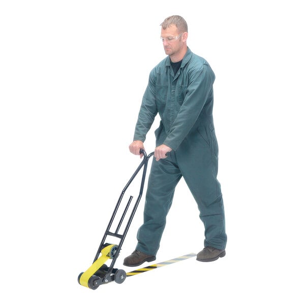 A man wearing a green jumpsuit uses a Vestil steel floor tape applicator to pull yellow tape.