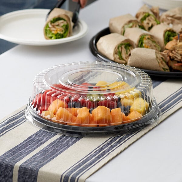 A Visions black plastic catering tray with a high dome lid holding fruit and a sandwich on a table outdoors.