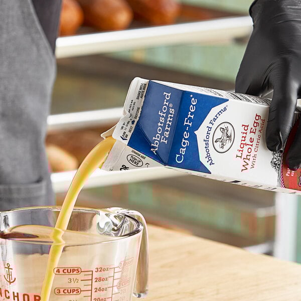 A person in black gloves pouring Abbotsford Farms liquid whole eggs into a measuring cup.