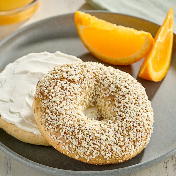 A plate with a bagel with white cream cheese and Hometown Provisions white sesame seeds with orange slices on the side.