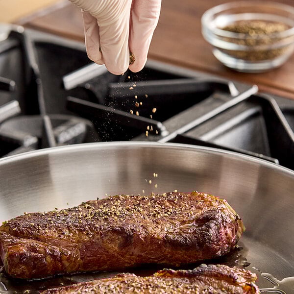 A hand sprinkling Hometown Provisions Coarse Grind Black Pepper on meat cooking in a pan.