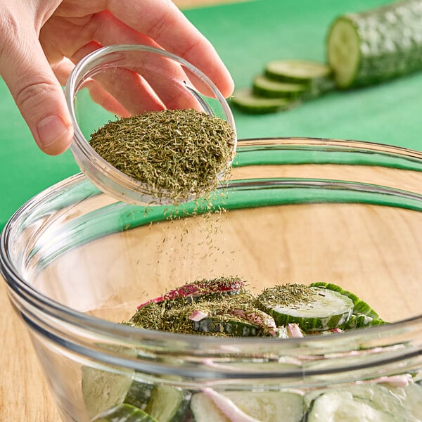 A hand pouring Hometown Provisions Dill Weed into a bowl of cucumbers.